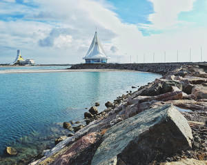 岩石碼頭上的現代帆形建築，旁邊是平靜的藍綠色海水。 — 5:4 壁紙