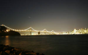 星空下，夜間城市燈光映照水面上的一座亮燈吊橋。 — 城市景觀 壁紙