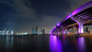一座紫色照亮的橋樑橫跨黑暗的水道，背景是城市天際線。 — 城市 壁紙