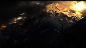 一座積雪的山峰被金色日出光照亮，背景是黑暗的暴風雲，下方是森林剪影。 — 簡約暗黑美學 壁紙