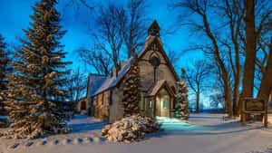 暮色中一座被雪覆蓋的舒適木造小教堂，周圍有光禿的樹木。 — 冬季風景 壁紙