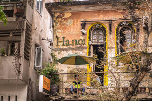 顧客坐在綠色遮陽傘下，位於一座褪色建築的咖啡館，建築有黃色拱形窗戶。 — 咖啡美學 壁紙