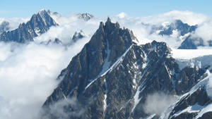 一座高聳的山峰在翻騰的雲層之上崛起，積雪附著在岩石斜坡上。 — 山景 壁紙