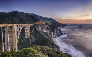夕陽下懸崖上的混凝土拱橋，波濤洶湧，群山聳立 — 日落 壁紙