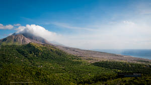火山在藍天下向綠色山丘和海洋噴出煙霧 — 山脈 壁紙