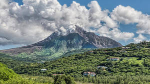 火山在藍天白雲下，於綠色山丘上冒煙，山丘上散落著房屋。 — 城市 壁紙