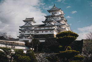 白色日本城堡在石山上，周圍有綠樹和部分多雲的天空 — 簡約暗黑美學 壁紙