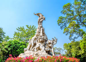 石山羊雕塑，周圍有綠樹和粉紅色花朵，背景是藍天。 — 廣州 壁紙