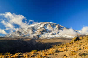 在清澈的藍天下，雲朵環繞著岩石山坡的雪山峰頂。 — 雪山 壁紙