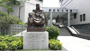 青銅雕像，一位坐著的成人抱著孩子，位於現代建築前，周圍有綠植。 — 廣州 壁紙