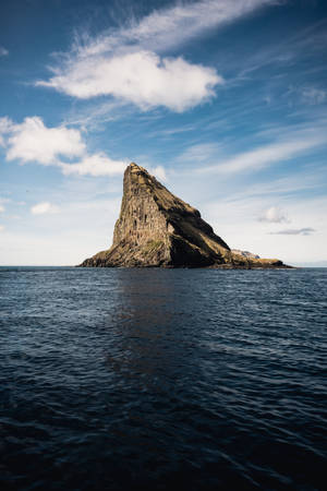 一座陡峭的岩石島嶼獨自矗立在開闊的海洋中，天空部分多雲。 — 法羅群島 壁紙