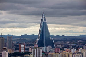 一座高大的玻璃金字塔建築在多雲的天空下矗立於城市之上。 — 地獄客棧 壁紙
