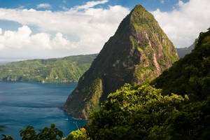 一座陡峭的綠色山峰俯瞰著藍色海洋，上方有零星雲朵。 — 山景 壁紙