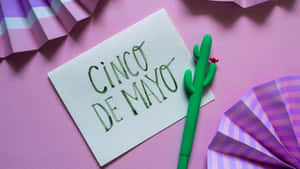 手寫 Cinco de Mayo 便條，配有綠色仙人掌筆和紫色紙扇，背景為粉紅色。 — 慶祝 壁紙