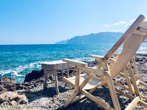岩石海岸上的木製沙灘椅和邊桌，背景有海洋和山脈。 — Chill 壁紙