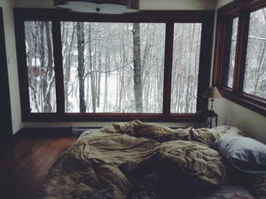臥室中未整理的床，透過大窗戶可看到雪森林景色。 — 冬季房屋 壁紙