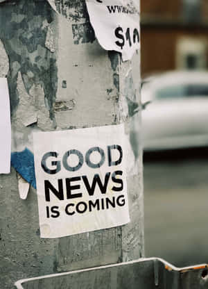 柱子上的一張破舊貼紙寫著「GOOD NEWS IS COMING」，處於城市雜亂之中。 — Kindle 壁紙