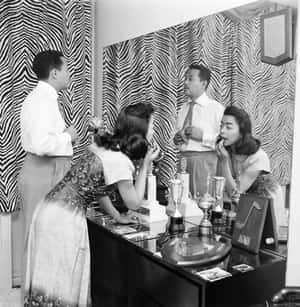 一男一女穿著1950年代服裝，在鏡子前化妝，梳妝台上放著獎盃。 — 喬爾·恩比德 壁紙