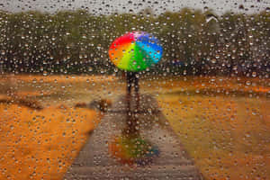 人拿著彩虹傘在濕路上行走，透過有雨痕的窗戶。 — 下雨 壁紙