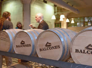 帶有 Balcones 標誌的木製威士忌桶，釀酒廠內人們在交談。 — 桌面 壁紙