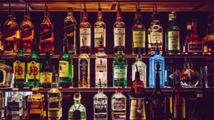 一排色彩繽紛的酒瓶在溫暖燈光下的吧台架上。 — 酒吧 壁紙