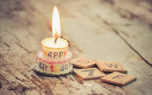一支有 'Happy Birthday' 字樣的生日蠟燭，在木頭上的 Scrabble 字母磚旁邊燃燒。 — 生日快樂蛋糕 壁紙