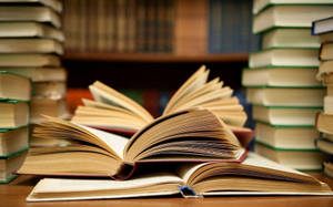 圖書館木桌上堆疊的書本圍繞著打開的書。 — 讀書 壁紙