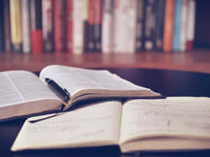 打開的書本和筆在筆記本上，前面是書架，適合學習。 — 閱讀 壁紙