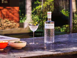 一杯琴酒馬丁尼配橄欖，旁邊放著一瓶 The Botanist，在戶外大理石吧檯上。 — 4:3 壁紙