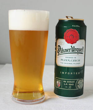 一杯Pilsner Urquell啤酒在白色表面上，旁邊是它的綠色罐子。 — Motorola Razr 70 Ultra 壁紙