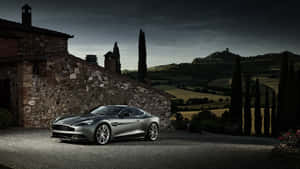 銀色 Aston Martin 停泊在石頭別墅旁，伴隨著柏樹，在暮色中。 — 奧斯頓馬丁 壁紙