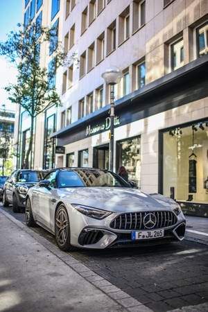 銀色 Mercedes-AMG GT 停泊在陽光明媚的城市街道上，旁邊是一家豪華精品店。 — Mercedes Amg iPhone 壁紙
