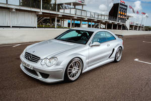銀色 Mercedes-Benz CLK GTR 賽車在賽道上，背景有看台和旗幟。 — Mercedes Benz C300 壁紙