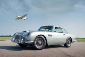 銀色經典Aston Martin DB5停在飛機跑道上，一架小飛機飛過頭頂。 — 奧斯頓馬丁 壁紙
