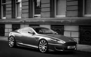 一架銀色Aston Martin DB9停泊在城市街道上，前面是石建築。 — Steam Deck 壁紙