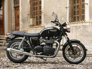 黑色 Triumph Bonneville 摩托車在鵝卵石街道上，靠近石頭建築 — 改裝摩托車 壁紙