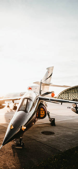 流線型戰鬥機在停機坪上沐浴在金色日出光芒中，靠近機庫。 — iPhone 15 壁紙