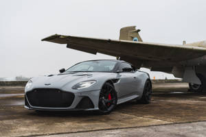 銀色Aston Martin跑車停泊在復古軍用飛機旁邊的停機坪上。 — 豪華車 壁紙