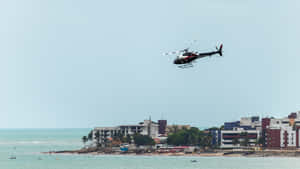 直升機飛越海洋和海灘小鎮的建築物 — 5120x2880 壁紙