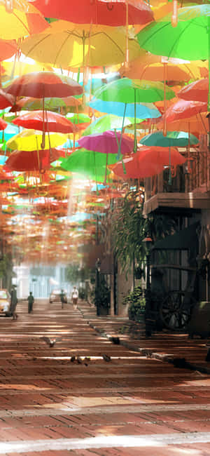 五彩繽紛的雨傘懸掛在頭頂，在有行人的磚砌街道上投下斑駁的光影。 — iPhone 16 Pro Max 壁紙