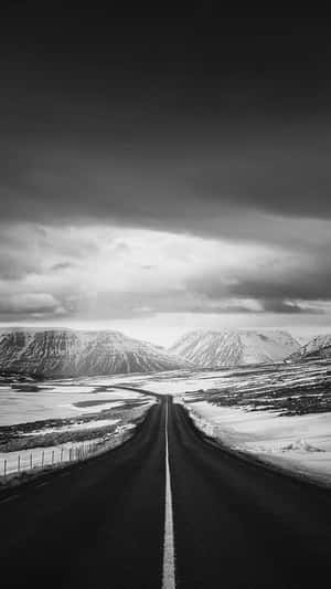 一條筆直的道路穿過積雪的山脈，天空多雲。 — 白色 iPhone 壁紙