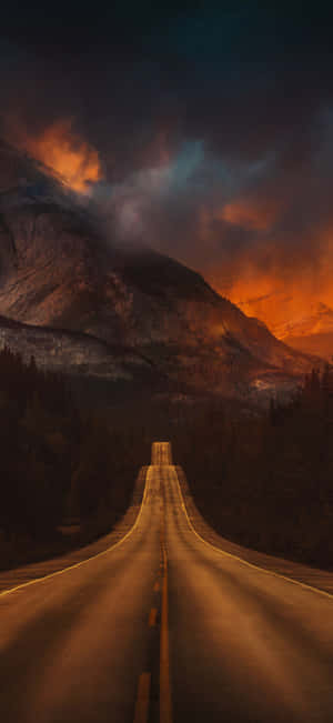 一條蜿蜒穿過森林的道路在戲劇性的暮色天空下通往群山。 — iPhone 18 壁紙