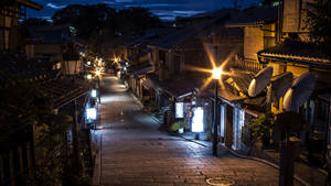 一條狹窄的石頭街道，兩旁是傳統的日本建築，夜晚被溫暖的燈籠照亮。 — 日式 壁紙