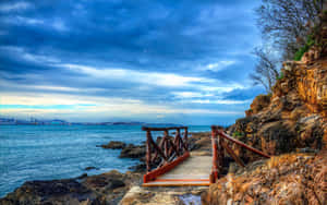 岩石懸崖上的木製步道，俯瞰海洋，遠處可見城市天際線。 — 2880x1800 壁紙