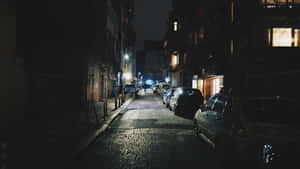 夜晚昏暗的鵝卵石小巷，停有汽車和發光的窗戶。 — 黑夜 壁紙