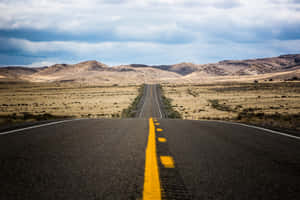 一條空蕩蕩的道路，帶有黃色中心線，穿過乾燥的景觀通往遠方的丘陵。 — 道路 壁紙
