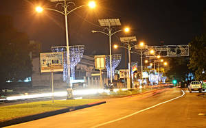 夜晚的城市街道，閃爍的路燈和節日裝飾，模糊的車燈光線劃過道路。 — 城市燈光 壁紙