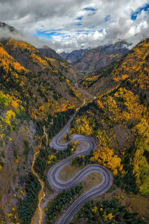 多雲天空下蜿蜒山路穿過秋林山谷 — 科羅拉多 壁紙