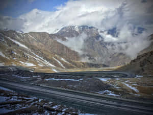 蜿蜒的山路，帶有雪斑和雲朵，背景是崎嶇的山峰。 — 道路 壁紙