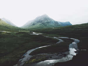 一條蜿蜒的河流流經綠色山谷，背景是霧濛濛的山脈。 — 山景 壁紙